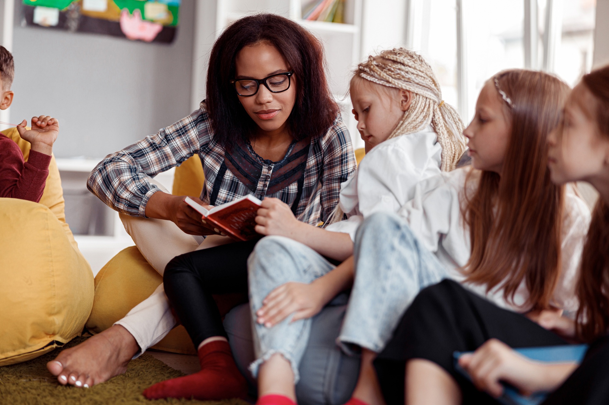 Teacher in glasses sitting on a pouf and reading for children in school